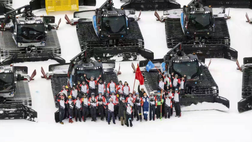 Aerial view of group of people waving, standing in front of rows of snow grooming machines