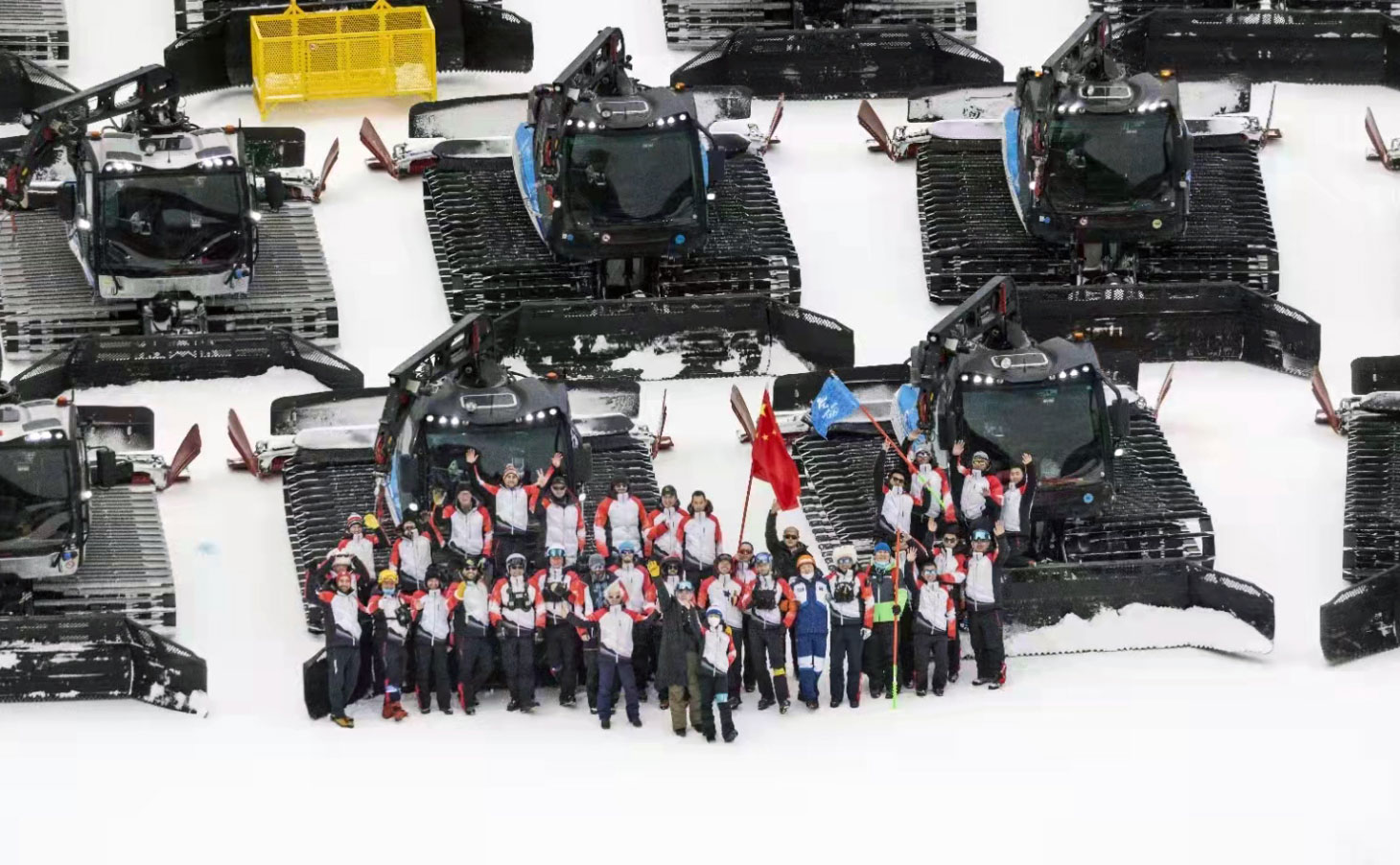 Aerial view of group of people waving, standing in front of rows of snow grooming machines