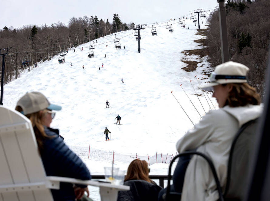 Two women seated in deck chairs with view of skiers and snowboarders coming down mountain