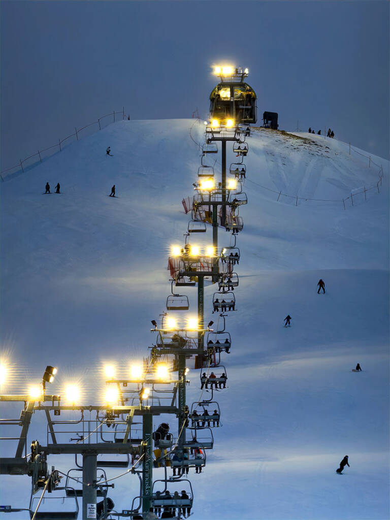 Calgary, Alberta, Canada. Dec 31, 2025. Night Skiing at WinSport Canada Olympic Park Under Bright Floodlights. Vertical shot of a glowing chairlift line ascending a snowy hill with skiers at dusk.