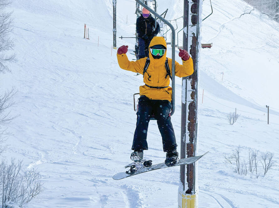 NISEKO, JAPAN, 2nd FEBRUARY 2024: Snowboarder strikes a playful pose on single chairlift above snowy slopes of Japanese ski resort. Brave tourist on iconic ski lift without any safety bar or ski rest.